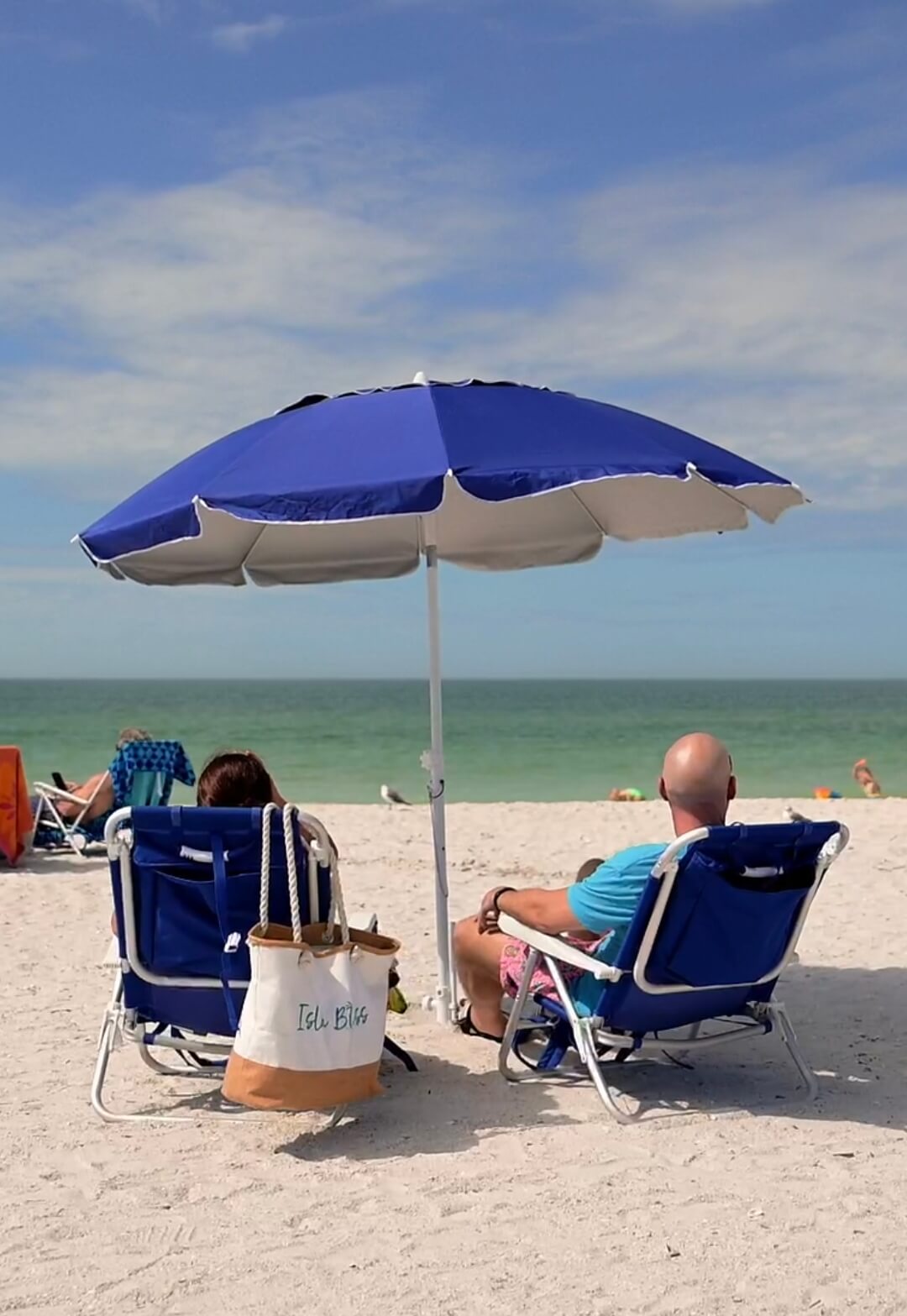 Couple relaxing on beach chairs under a blue umbrella facing the ocean