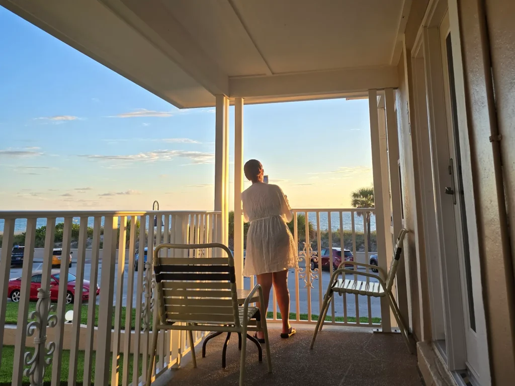 A woman in a white dress stands on a covered balcony, looking out at the ocean during sunset