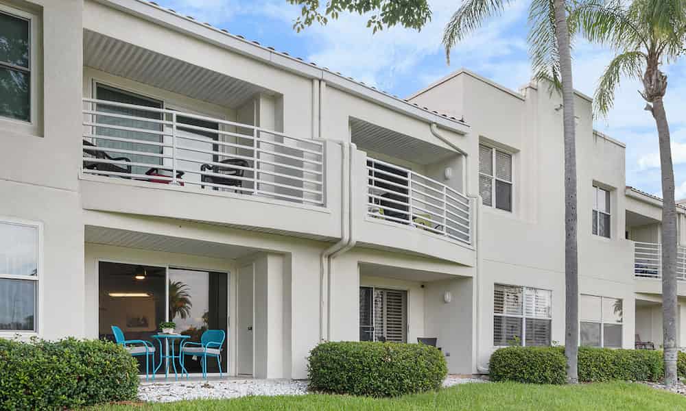 Modern two-story apartment building with balconies, patio seating, and palm trees in a landscaped outdoor area