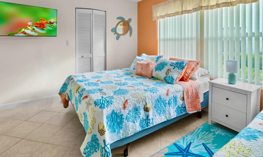 Bright coastal-style bedroom with coral-themed bedding, white nightstand, and large window with vertical blinds letting in natural light