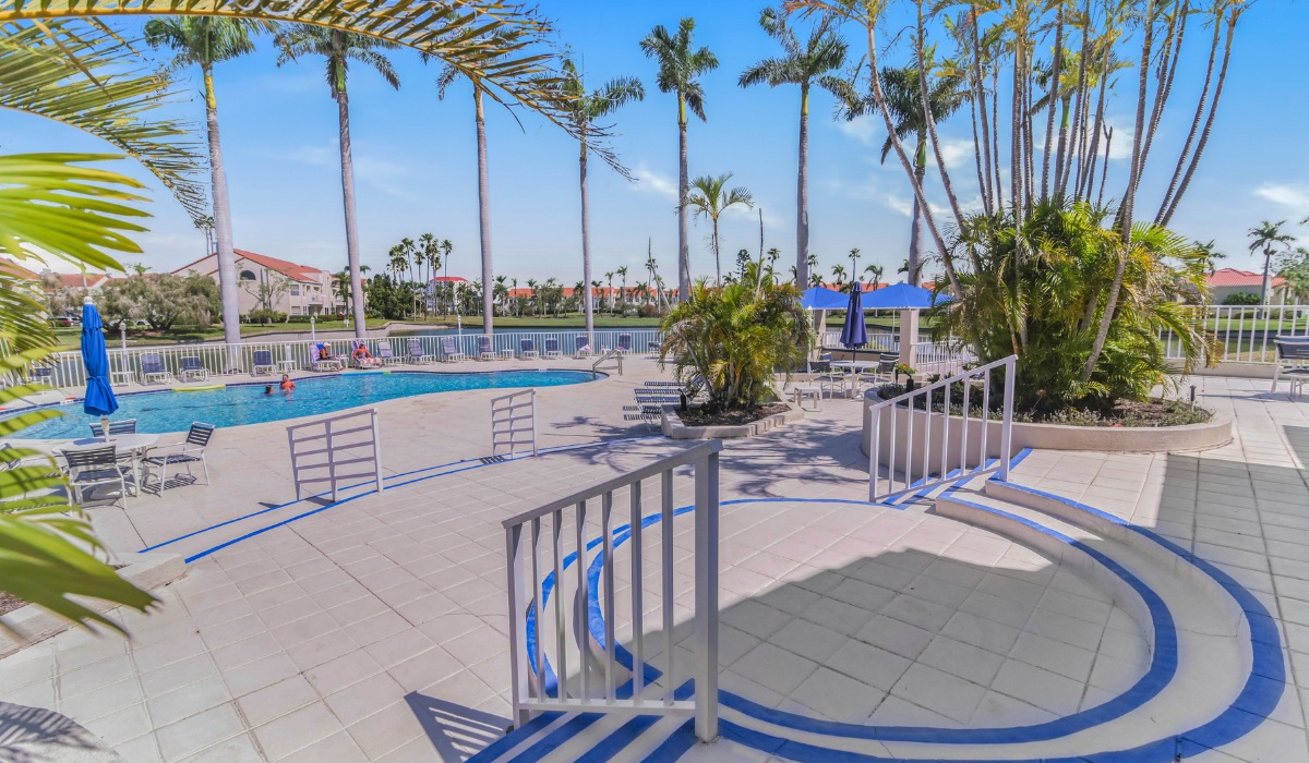 A sunny outdoor resort pool deck area with palm trees, lounge chairs, a swimming pool, and tiled stairs leading down to the patio